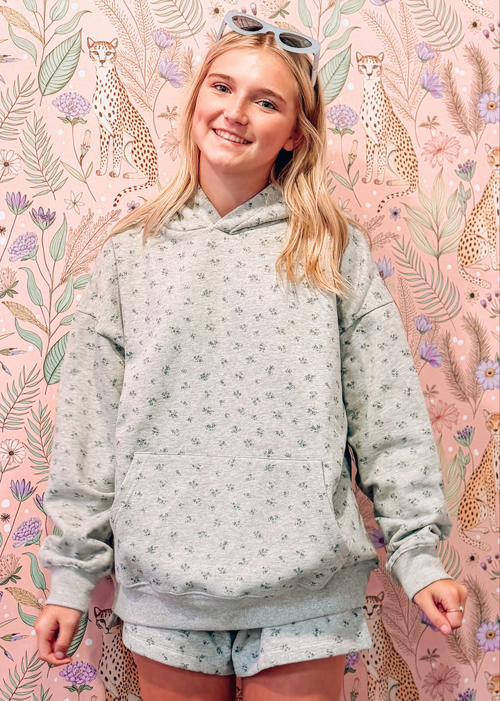 A young woman stands in front of a pink floral and animal print backdrop, wearing a heather grey patterned hoodie and matching shorts.