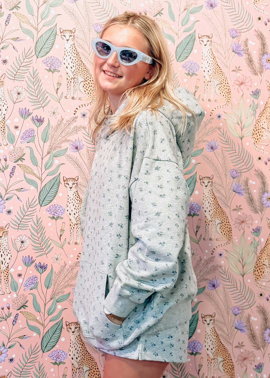 A young woman with blonde hair and blue-tinted sunglasses models a light blue patterned hoodie and matching shorts set against a pink floral wallpaper with cheetah motifs.
