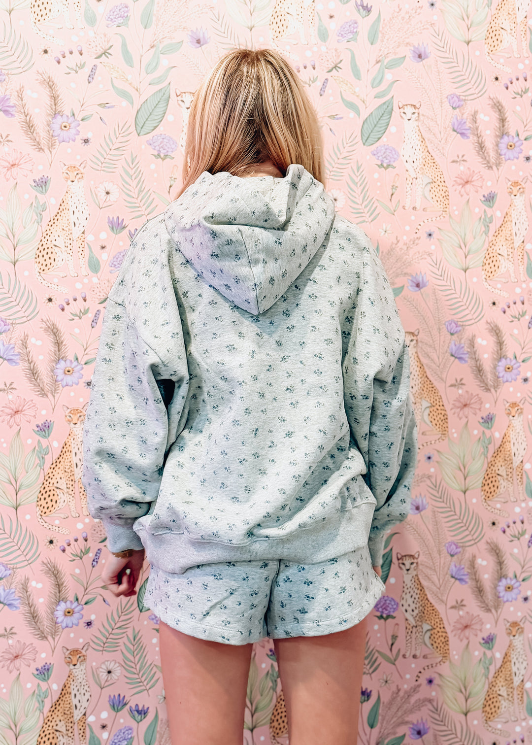 A woman viewed from behind wearing a light sage green and white floral print hoodie and matching shorts, standing in front of a pink floral wallpaper background.