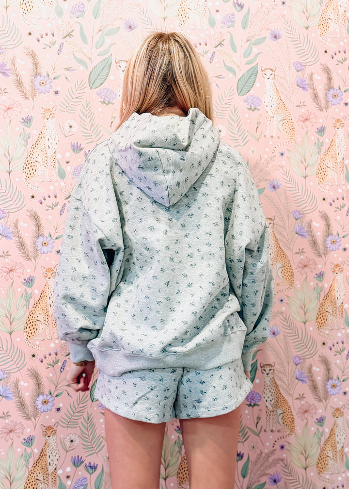 A woman viewed from behind wearing a light sage green and white floral print hoodie and matching shorts, standing in front of a pink floral wallpaper background.