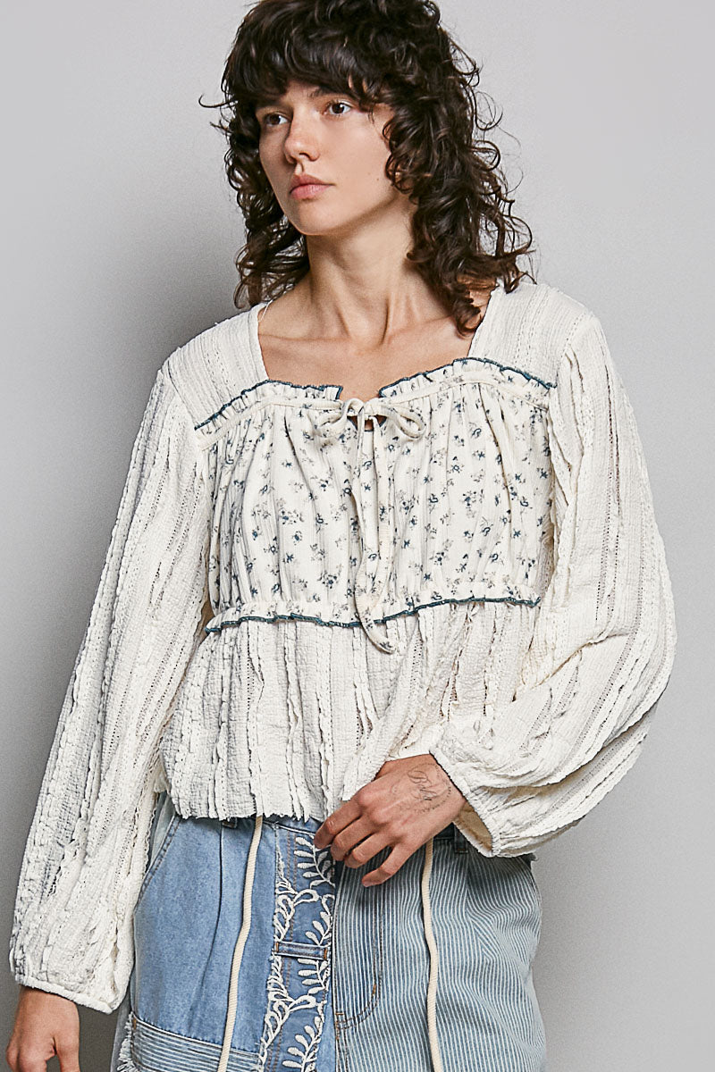 Woman wearing a white blouse with floral patterns and blue jeans against a gray background