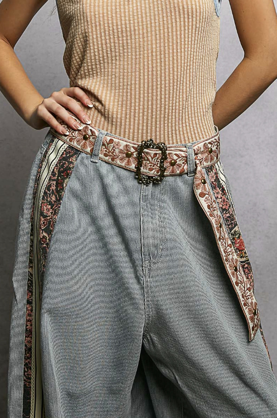 Person wearing a beige ribbed tank top and gray pants with decorative belt and side panels against a gray background