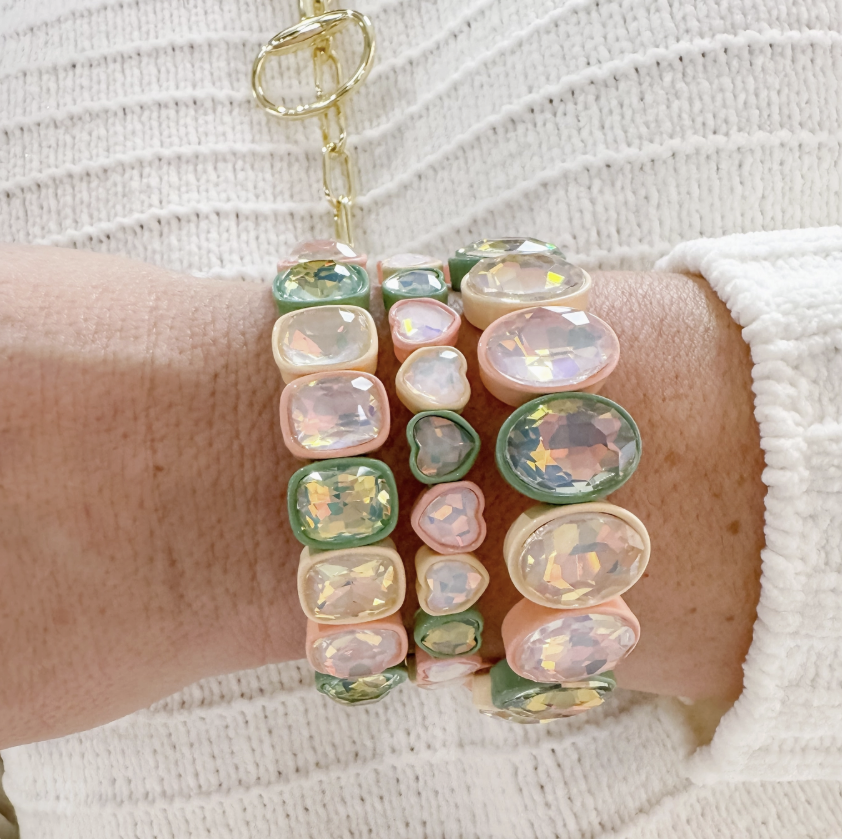 A close-up of a person's wrist wearing a stack of pastel gemstone bracelets, including pink, green, and iridescent oval and heart-shaped stones, with a gold clasp bracelet partially visible above.