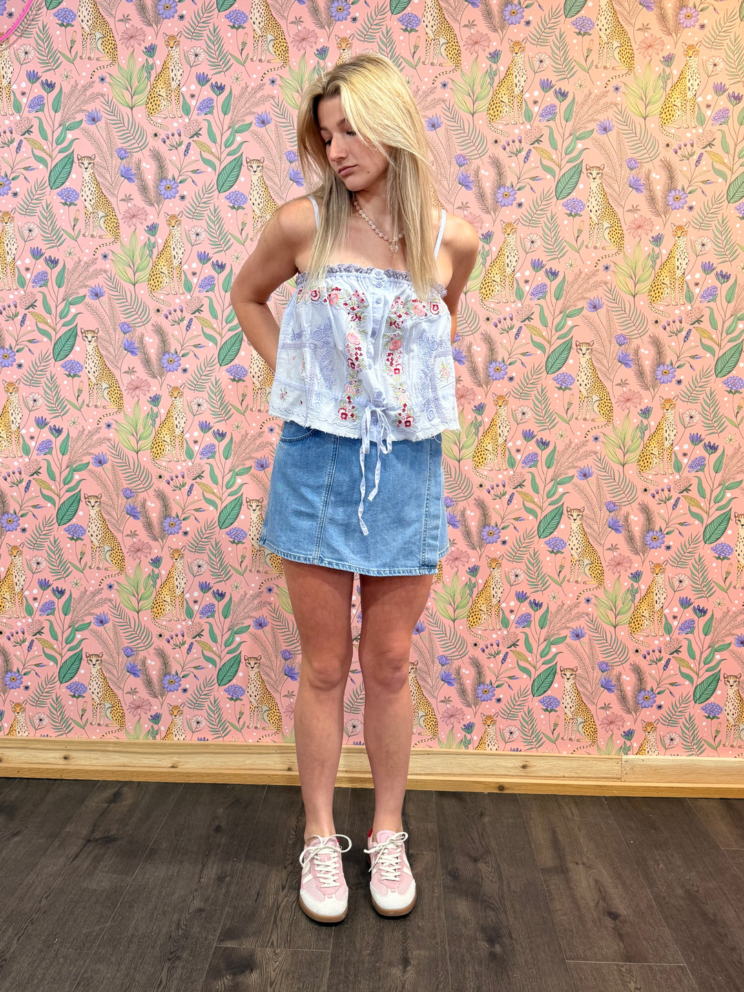 Woman standing in front of a colorful patterned wall with a floral top and denim skirt.