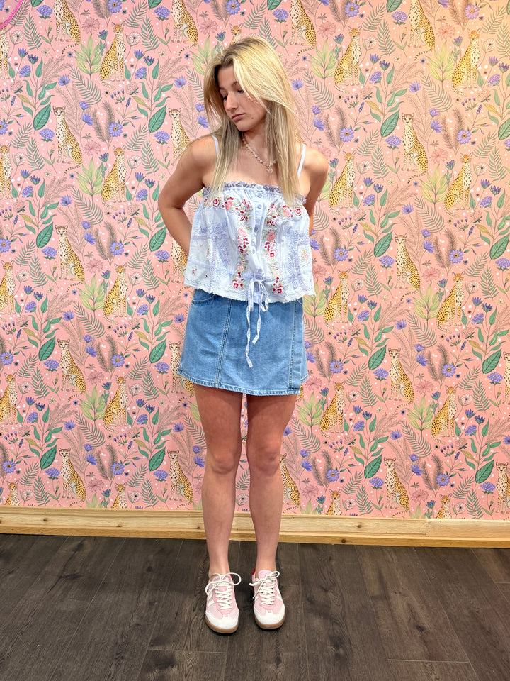 Woman standing in front of a colorful patterned wall with a floral top and denim skirt.