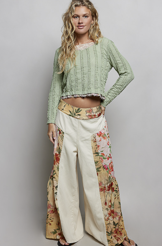 A woman modeling a light sage green cropped knit top with a lace neckline and wide-leg cream pants featuring floral side panels, against a plain light gray studio background.
