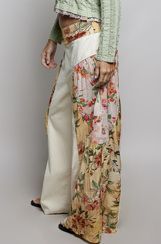 A person wearing wide-leg draped pants with a cream satin side and a floral printed side in beige, pink, and green, paired with a light green textured top against a plain gray studio background.