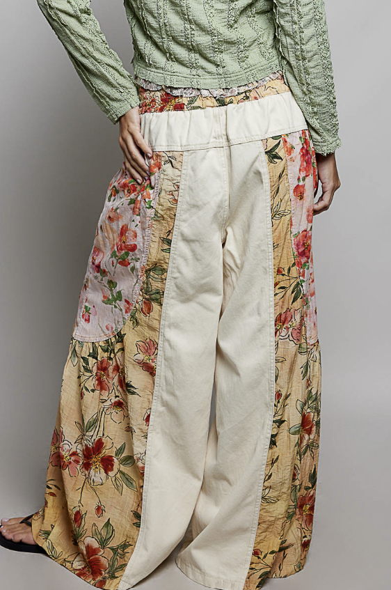 A person wearing wide-leg patchwork pants with cream panels and floral printed sections, paired with a light green textured top, shown against a plain gray studio background.
