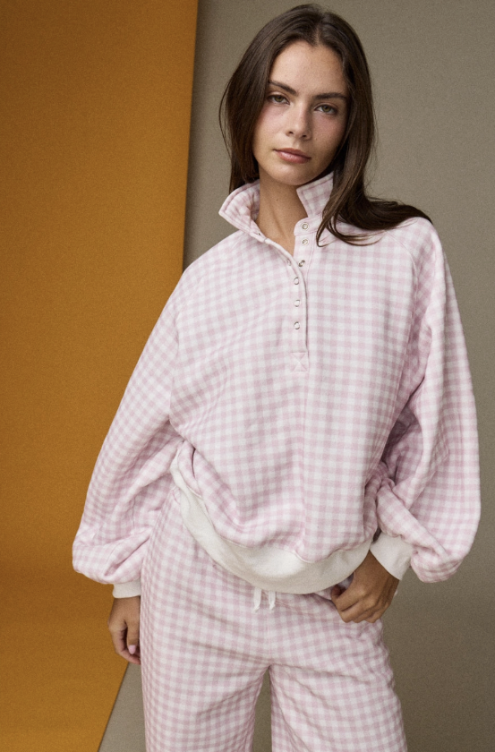 A woman wearing a light pink and white gingham pajama set with a half-button placket and matching pants, posed against a split orange and beige background.