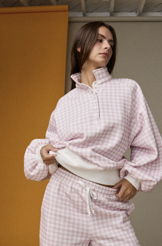 A woman wearing a light pink and white gingham check loungewear set, featuring a half-zip mock neck sweatshirt and matching drawstring pants, posed against a split orange and gray background.