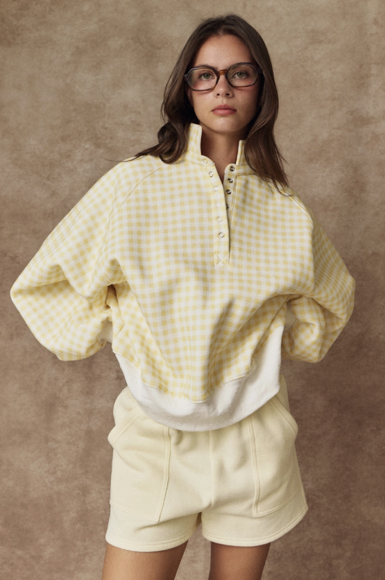 A woman wearing a pale yellow and white gingham quarter-zip pullover with a white ribbed hem and matching cream shorts, standing against a textured brown studio backdrop.