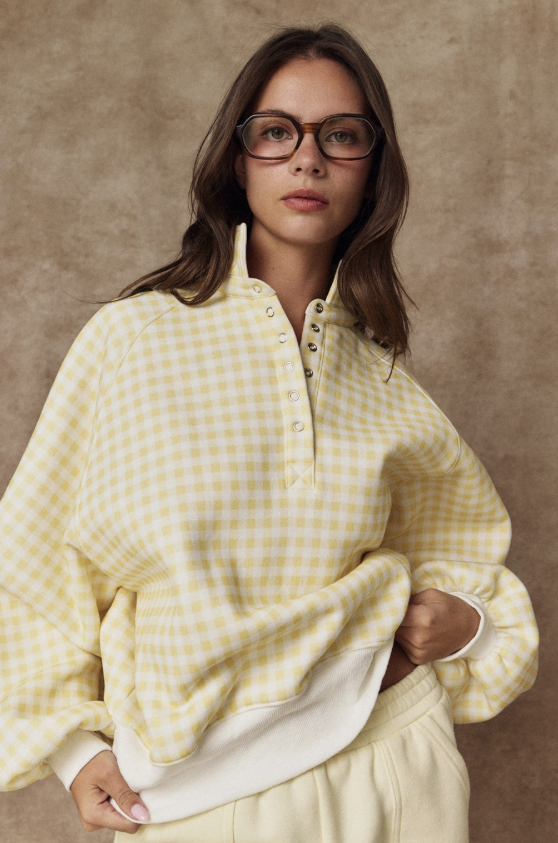 A woman wearing a pale yellow and white gingham quarter-placket pullover sweatshirt with a white ribbed hem, paired with cream sweatpants, set against a textured brown studio backdrop.