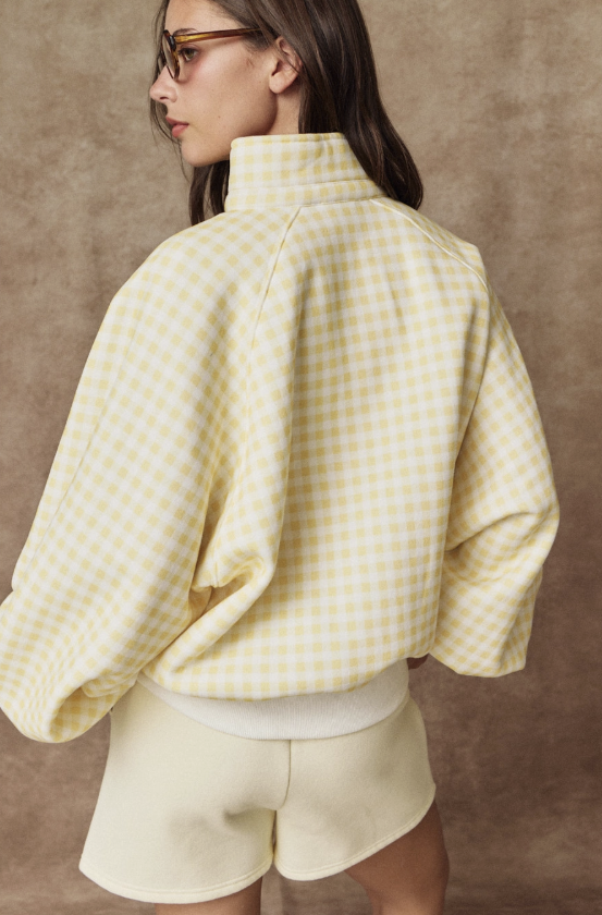 A woman shown from the back wearing a pale yellow and white gingham cropped jacket with a high mock neck and oversized sleeves, paired with matching cream shorts, against a textured brown studio backdrop.