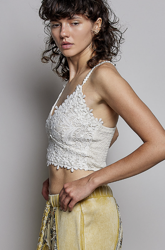 A woman modeling a white lace halter crop top paired with yellow satin pants against a plain gray studio background.