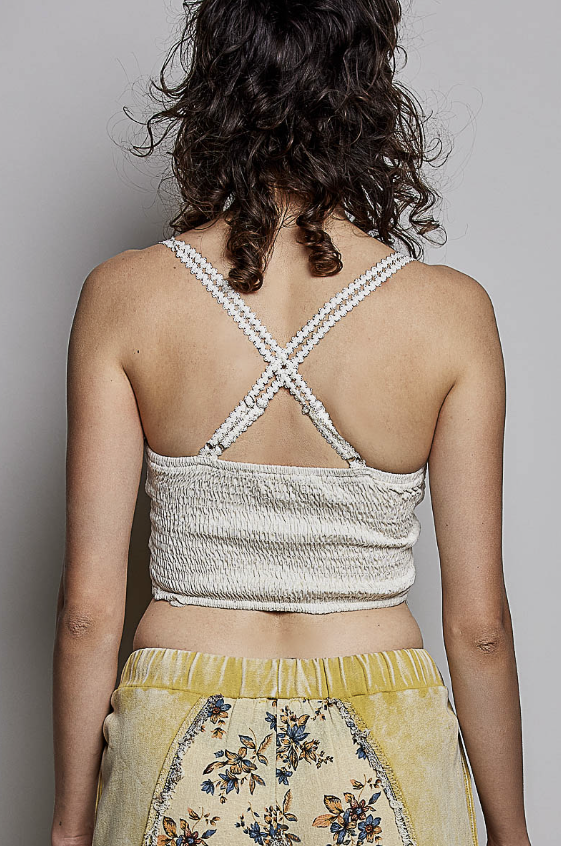 Back view of a woman wearing a textured cream-colored crop top with decorative braided straps and a yellow floral skirt against a plain gray background.