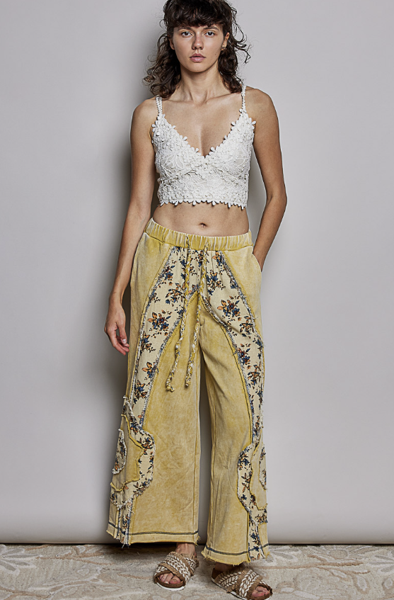 A model stands against a light gray studio wall wearing a white lace crop top and wide-leg yellow pants with floral print and tie details, paired with brown sandals.