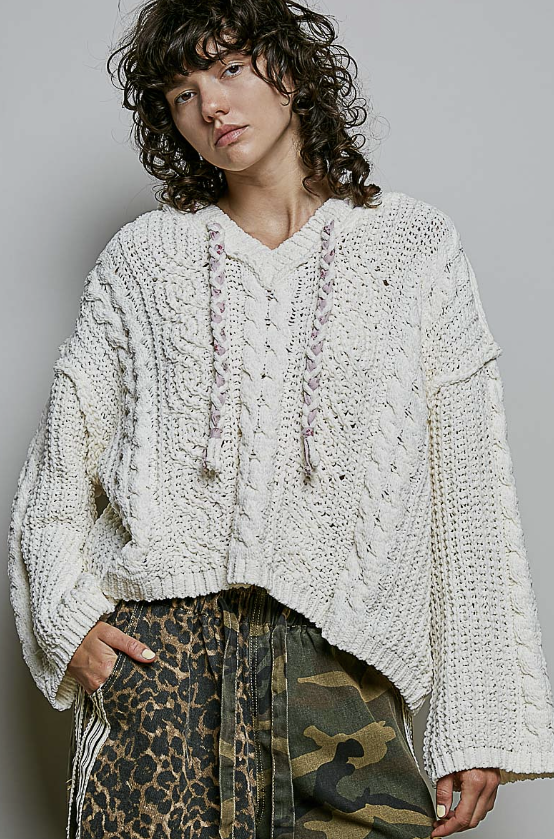 A model wearing an oversized, chunky cream-colored cable-knit pullover sweater with drawstrings, paired with leopard and camouflage print pants, against a plain gray studio background.