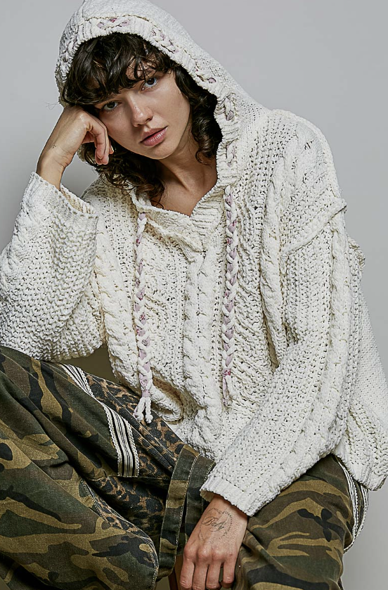 A model wearing a chunky cream-colored cable-knit hooded sweater and camouflage pants, posing against a neutral gray background.