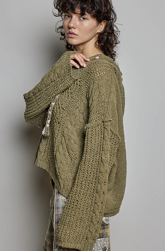 A woman wearing an oversized chunky-knit olive green sweater, photographed against a plain light gray studio background.