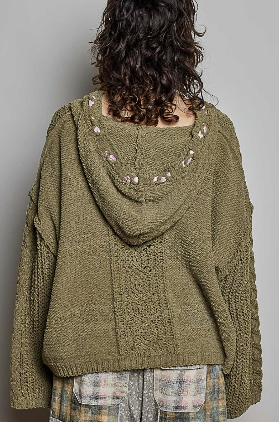 A person shown from the back wearing an olive green chunky knit hooded sweater with decorative floral embroidery around the neckline, paired with plaid pants, against a plain gray background.