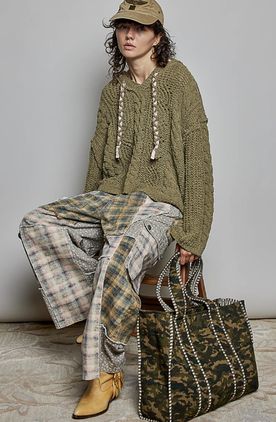 A model is seated on a wooden stool wearing an olive green chunky knit sweater, a beige cap, and a patchwork plaid shirt. They are holding a large green camouflage tote bag with pearl-like beaded handles against a neutral gray studio background.