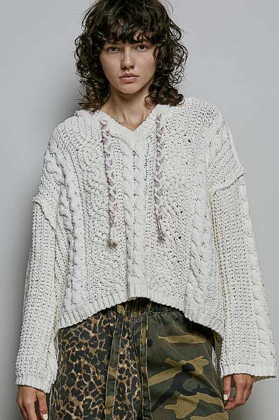 A model wearing a chunky, off-white cable-knit cropped sweater with drawstrings, paired with leopard and camouflage print pants, against a plain gray studio background.