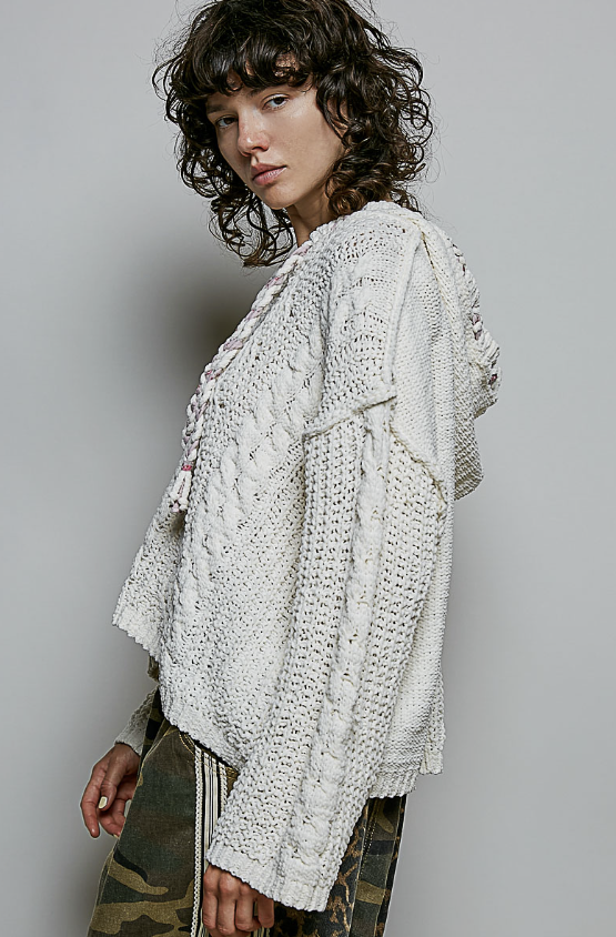 A model wearing a chunky, textured off-white knit cardigan with a hood, paired with camouflage pants, photographed against a solid gray studio background.