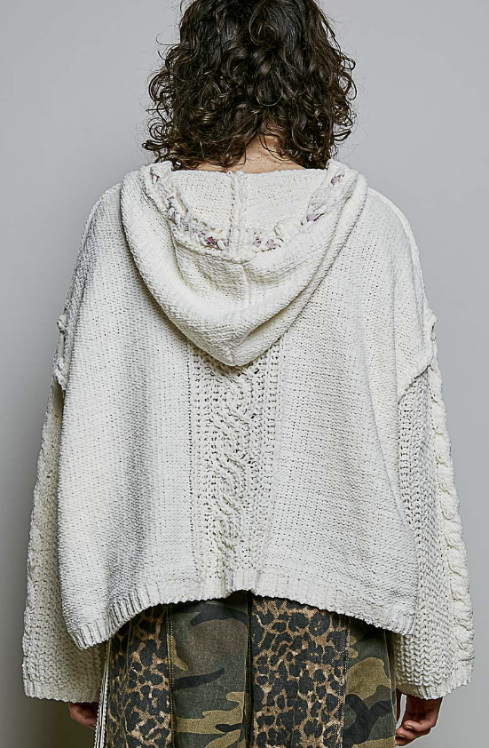 A person shown from the back wearing a chunky knit hooded sweater in an off-white/cream color, paired with camouflage and leopard-print pants, against a plain light gray studio background.