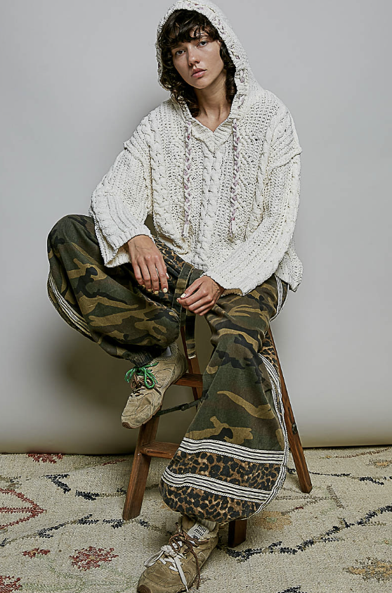 A model sits on a wooden stool wearing a chunky cream cable-knit hooded sweater, wide-leg camouflage cargo pants with mixed pattern panels, and beige lace-up boots against a neutral studio backdrop.