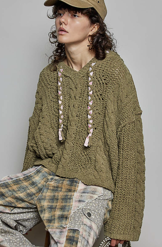 A model wearing an oversized olive green chunky-knit pullover sweater with pink braided drawstrings, paired with a tan cap and patterned plaid pants, set against a plain light gray studio background.