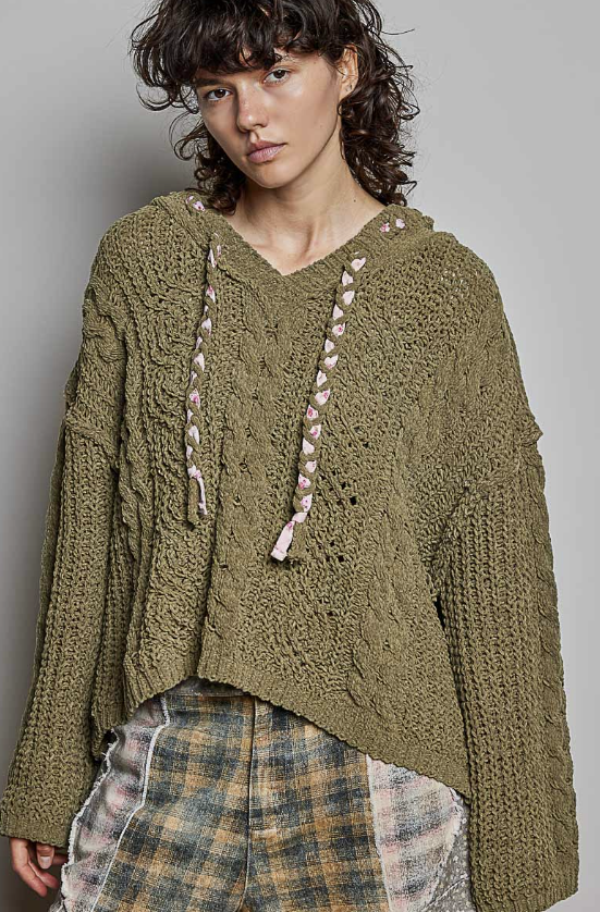 A model wearing an oversized olive green chunky knit hoodie sweater with pink braided drawstrings, paired with a plaid skirt, against a solid gray studio background.
