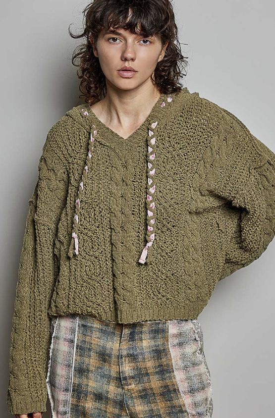 A model wearing a chunky olive green cable-knit cropped hoodie with pink and white braided drawstrings, paired with a plaid shirt and pants, against a plain gray background.