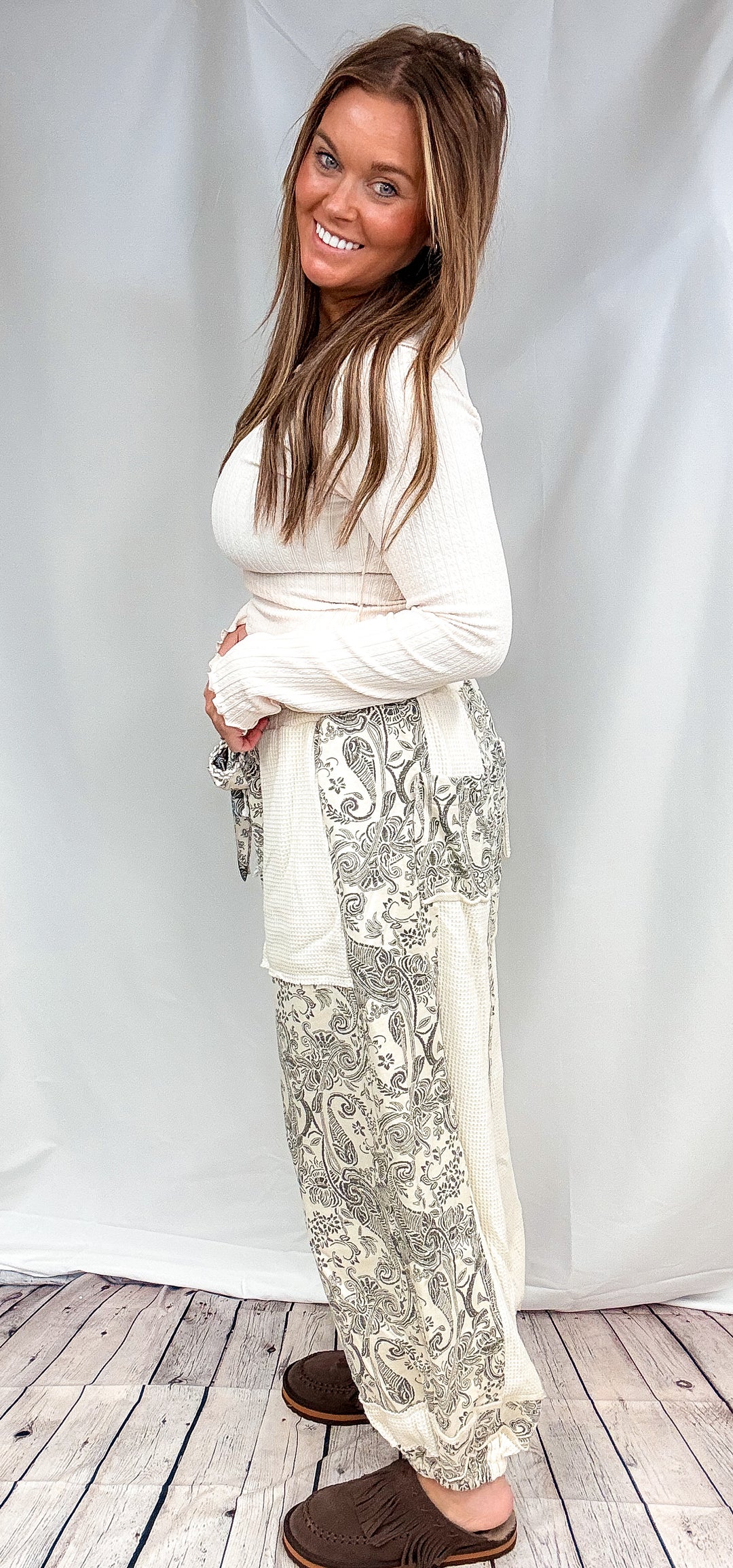 A woman modeling a cream long-sleeve top and matching cream pants with a gray paisley pattern, standing against a white studio backdrop on a light wood floor.