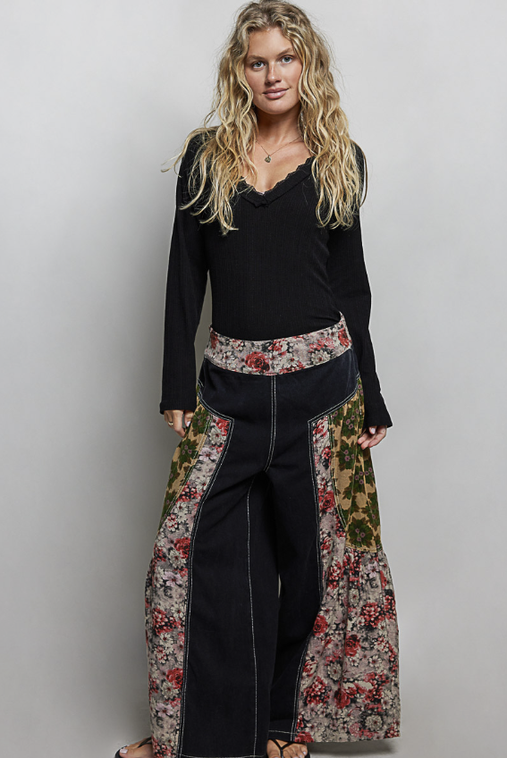 A woman modeling a black long-sleeve top with wide-leg pants featuring a black base and floral side panels in red, green, and beige tones, against a plain light gray studio background.
