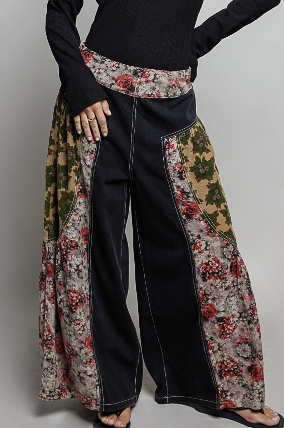 A person wearing wide-leg patchwork pants with a black main body and floral side panels, paired with a black long-sleeve top and black sandals against a plain light gray studio background.