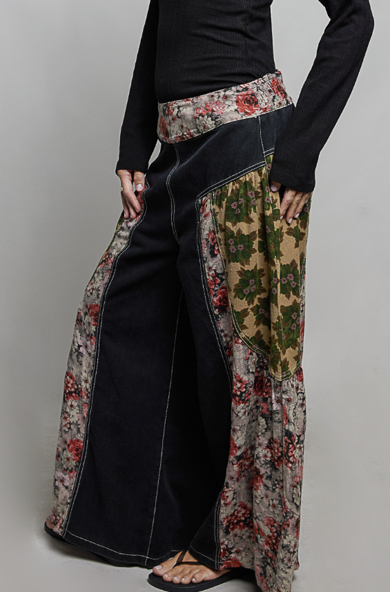 A person wearing wide-leg patchwork pants with dark navy denim panels and floral printed side panels in beige, red, and green, paired with a black long-sleeve top and black sandals against a light gray studio background.