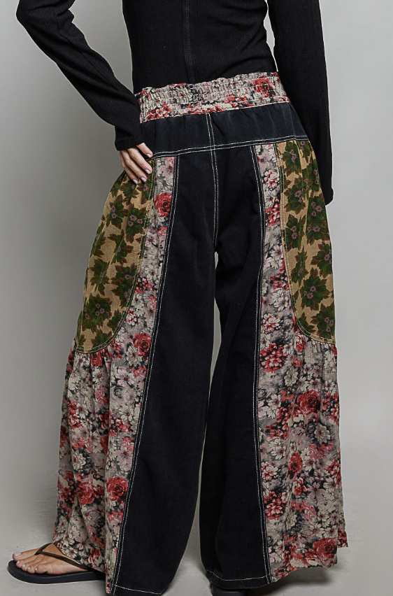 A person wearing wide-leg patchwork pants with dark charcoal denim-like panels and floral printed side panels in beige, green, red, and pink, paired with a black long-sleeve top and sandals against a light gray studio background.