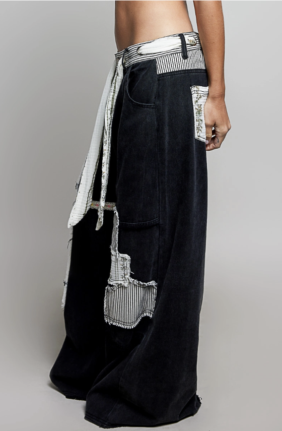 A person wearing very wide-leg black denim pants with contrasting white striped fabric panels, a striped waist tie, and frayed patch pockets, photographed against a light gray studio background.