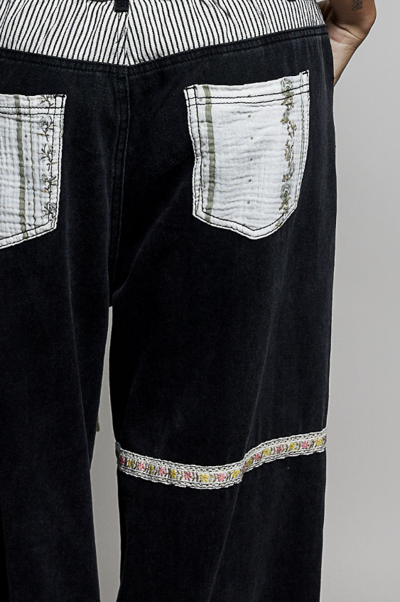 Close-up of the back of black corduroy pants featuring white patterned patch pockets and a decorative embroidered band near the hem.