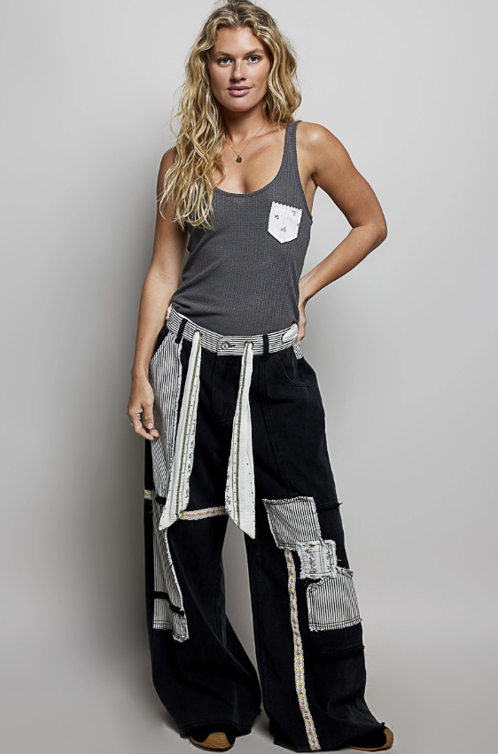 A woman modeling a dark gray ribbed tank top with a white chest pocket and black wide-leg pants featuring silver-gray patchwork fabric panels and a matching patterned belt, set against a plain light gray studio background.