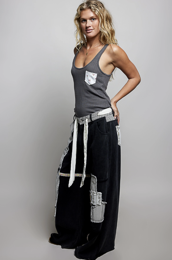 A woman modeling a dark gray sleeveless tank top with a white chest pocket and black wide-leg pants featuring white striped and textured side panels, against a light gray studio background.