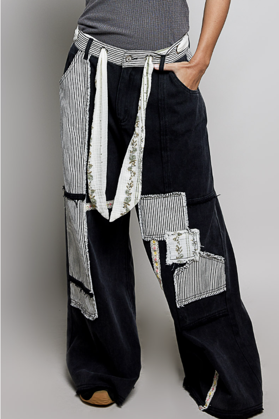 A person wearing very wide-leg black pants with large patchwork details made from white striped and patterned fabric panels, plus matching striped tie-waist cords, shown against a light gray studio background.