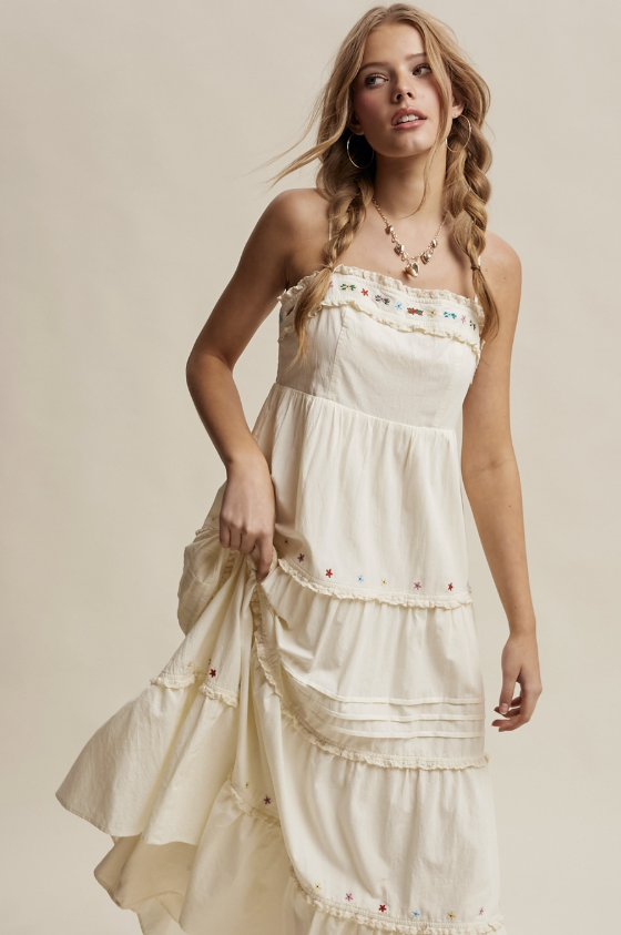 A woman modeling a cream-colored, tiered maxi dress with delicate ruffles and small colorful embroidered details. The dress has a smocked bodice and is styled with a gold necklace against a solid beige studio background.