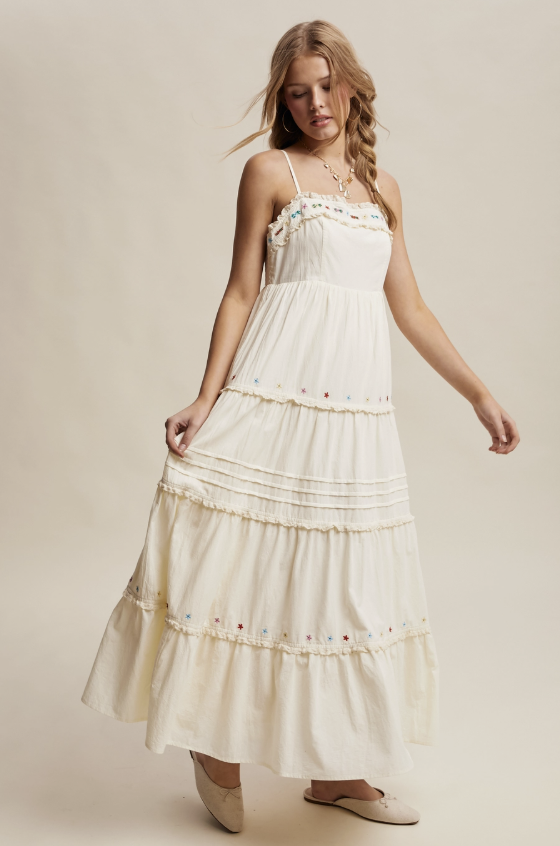 A woman modeling a long cream maxi dress with thin straps, delicate floral embroidery, and tiered ruffles against a solid beige studio background.