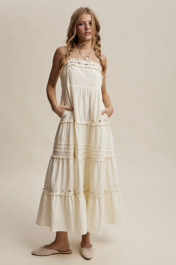 A woman modeling a tiered cream maxi dress with embroidered details and side pockets, paired with beige flat shoes, against a solid beige studio background.
