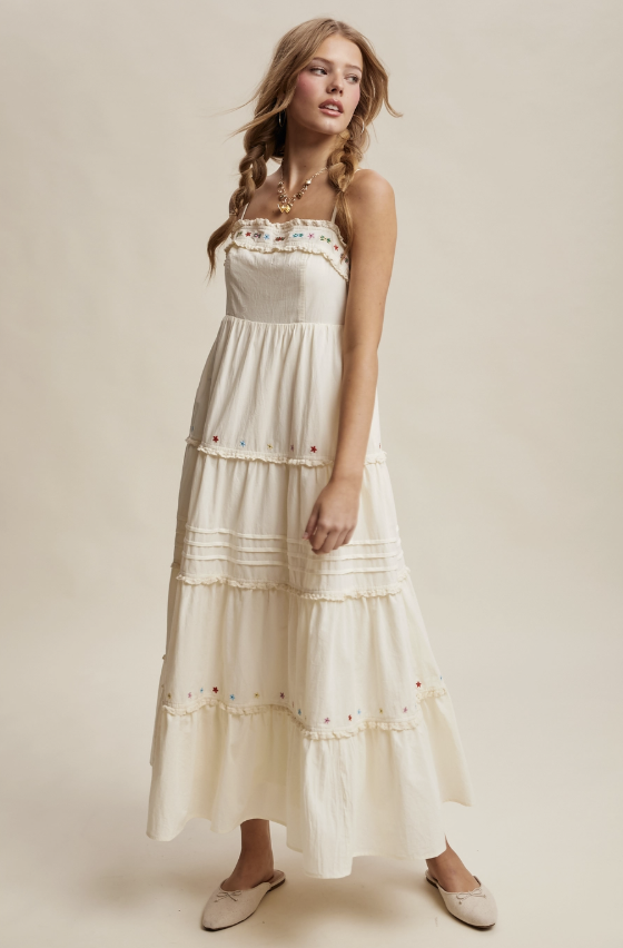 A woman modeling a cream-colored tiered maxi dress with delicate floral embroidery and ruffle trim, paired with beige flat shoes against a solid beige studio background.