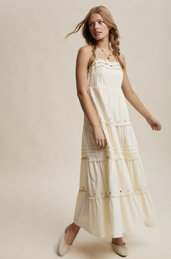 A woman modeling a cream-colored, tiered maxi dress with delicate embroidered details along the straps and skirt, paired with beige flat shoes against a solid beige studio background.