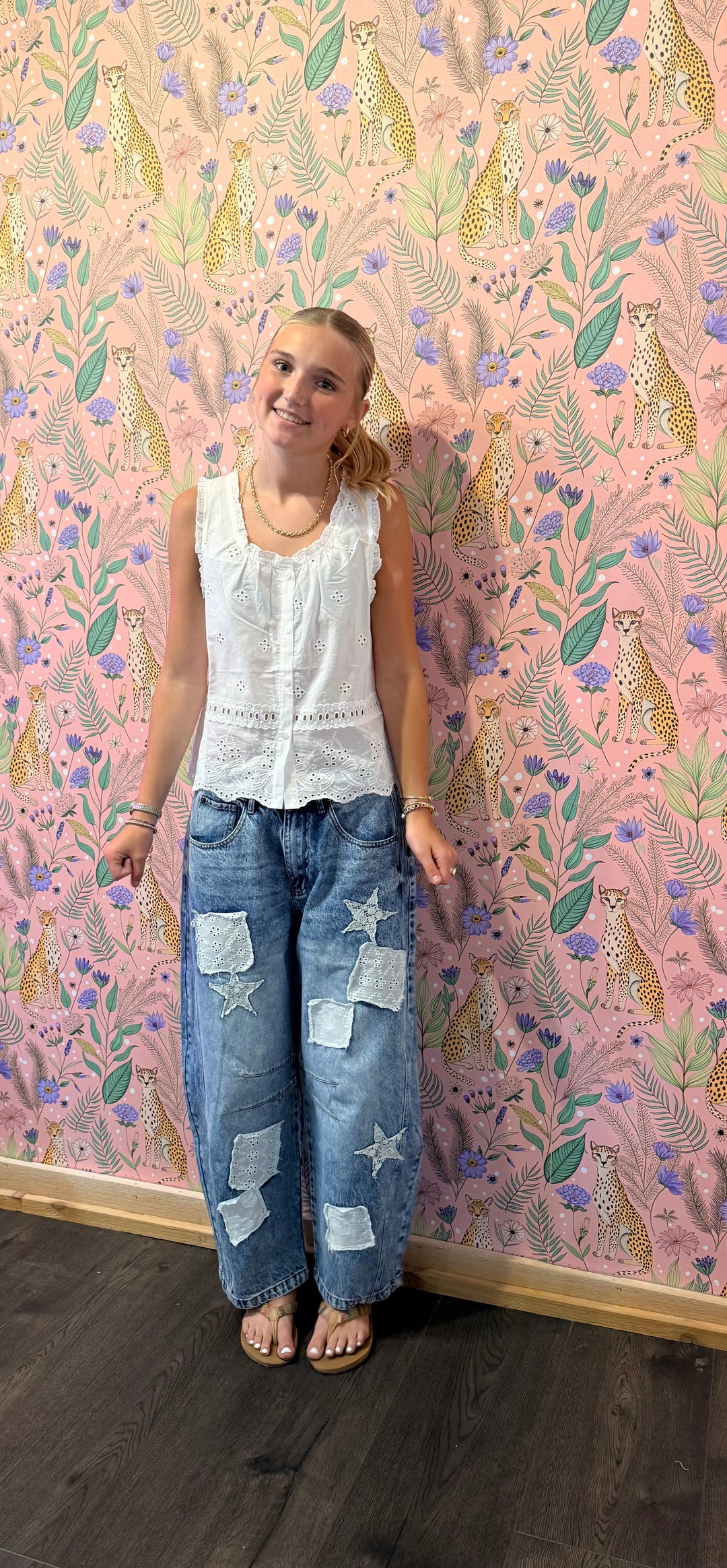 A woman stands against a pink patterned wallpaper background wearing a white sleeveless top and blue patchwork jeans with star patches.