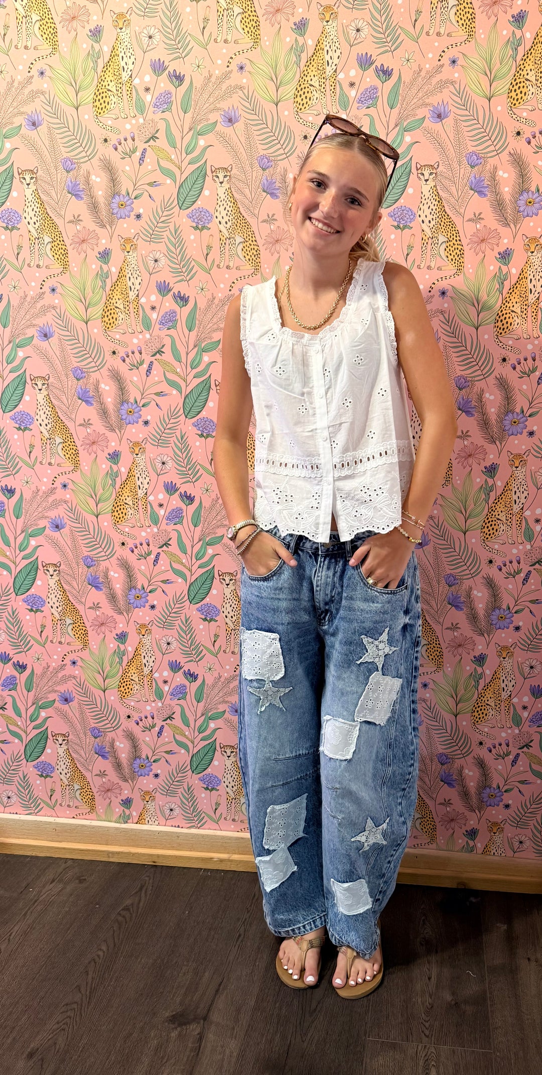 A woman stands in front of a pink leopard-print wallpaper wall, wearing a white sleeveless lace-trim top and blue patchwork distressed jeans with star patches.