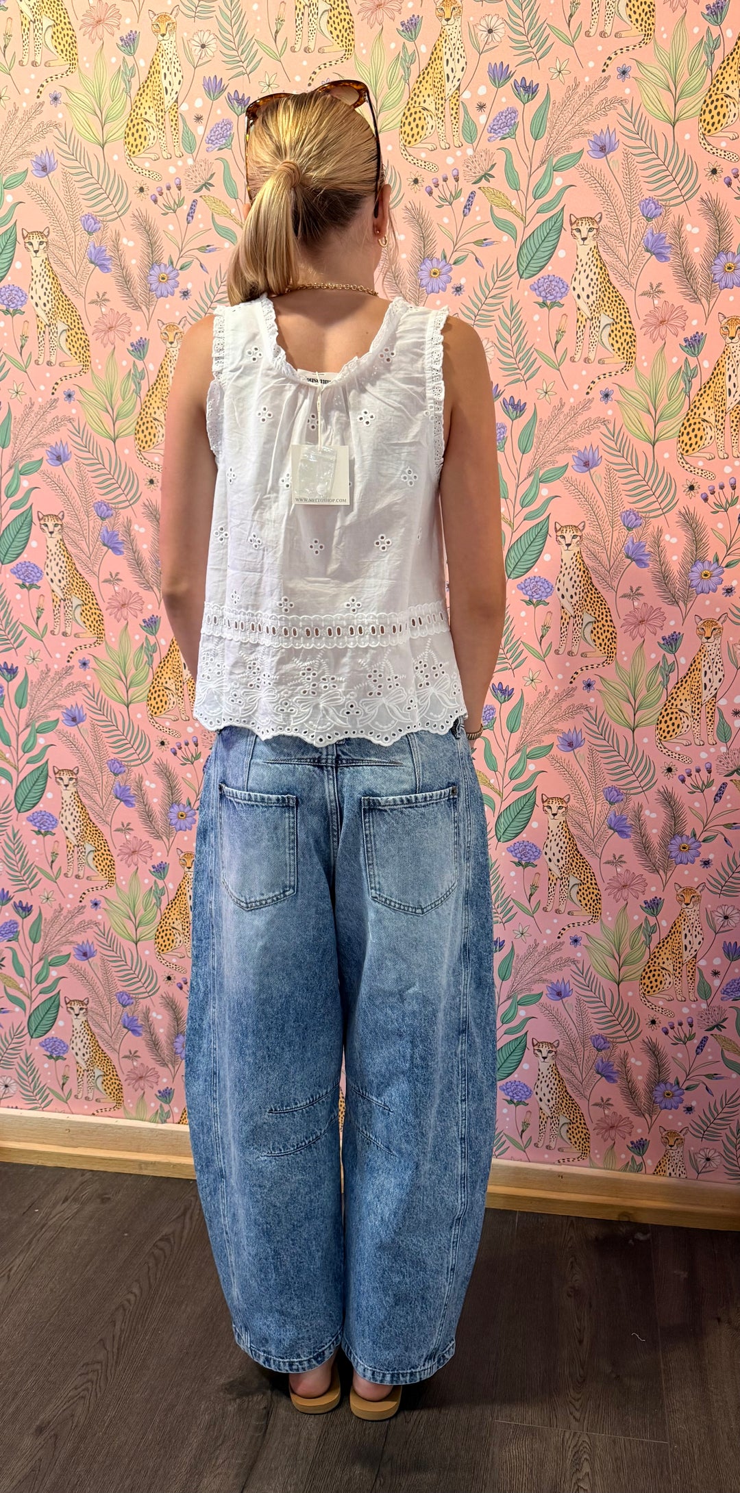 A woman stands with her back to the camera wearing a white sleeveless lace-trim top and wide-leg light blue jeans, standing in front of a pink patterned wallpaper background.